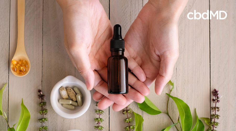 Someone holding a bottle of cbd in their hand with varoius cbd capsules and cbd sofgels around with some plants near by