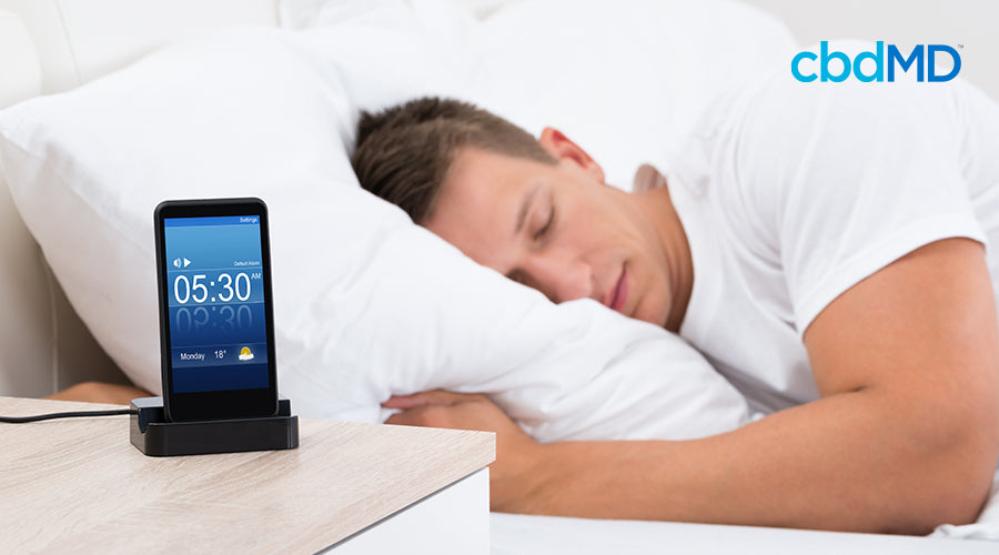 A man sleeping in a bed with his phone alarm sitting on a nightstand set for 5:30 am