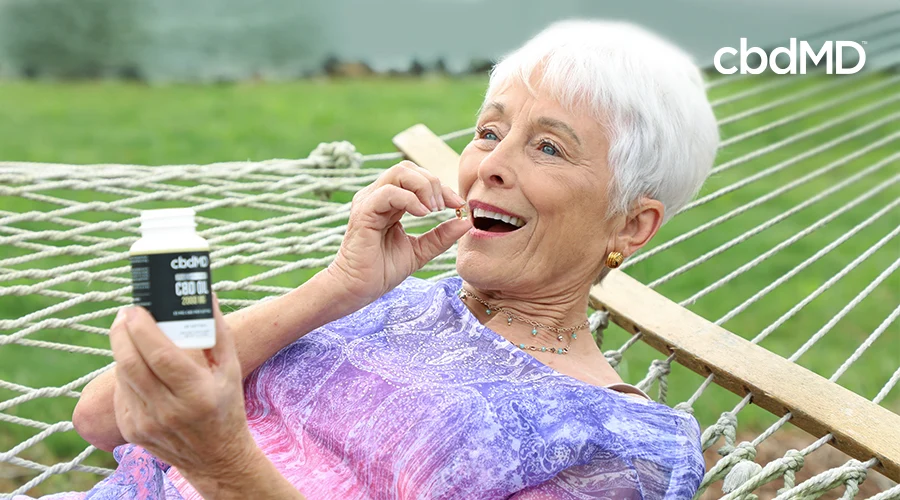 A woman laying on a hammock putting a cbd oil softgel in her mouth from a bottle of cbdMD full spectrum cbd oil 2000 mg softgels that she is holding in her hand  