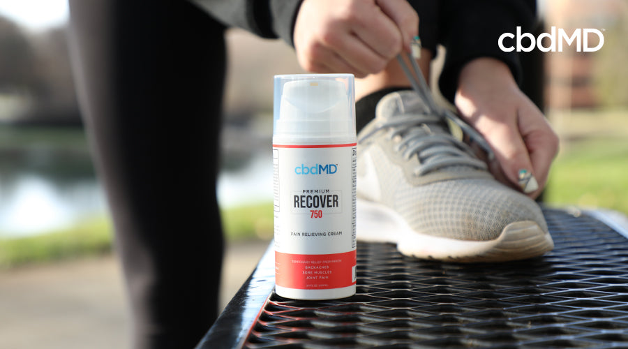 A woman with her shoe up on a bench while she ties it with a cbdMD recover 750 mg pain relieving cream container on the bench in front of her shoe