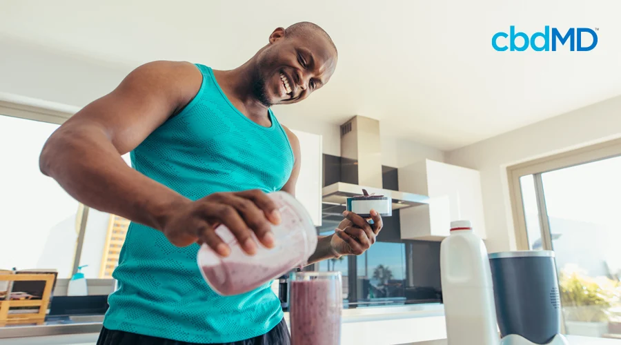 A man in a green tank top smiles as he makes himself an early morning protein shake