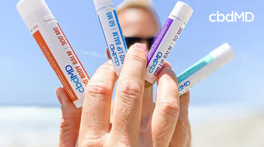 A man on the beach holds his hand out into the foreground of the image with an assortment of cbdmd cbd lip balm between his fingers