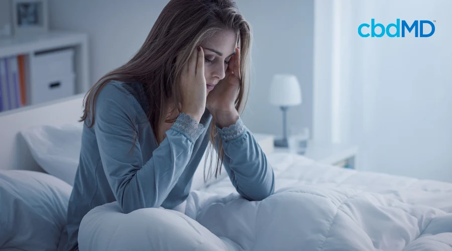 An attractive woman sits up in bed and holds her head with her eyes closed