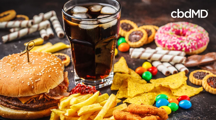 A table is laid out with a variety of junk food, including a glass of soft drink, chips, fries, candy, and a hamburger