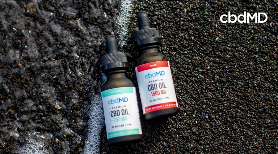 Bottle of cbdMD 1500 mg mint CBD oil tincture lays next to bottle of 750 mg orange CBD oil tincture by cbdMD on black sand with water next to bottles