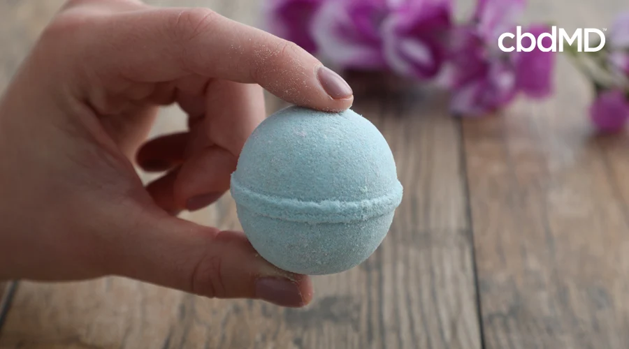 Manicured hand holds single eucalyptus Rejuvenation CBD bath bomb above wooden table with purple flowers in background