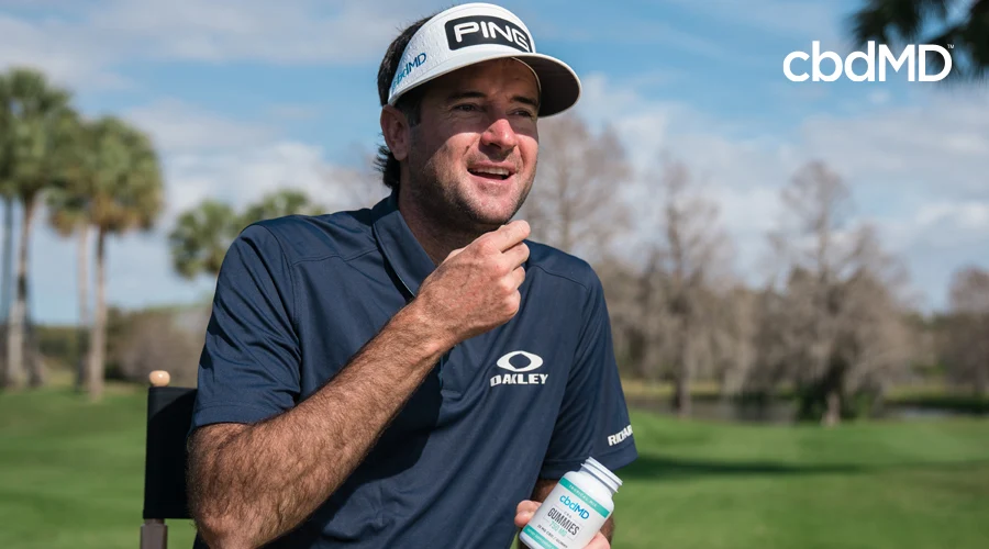 Masters Champion Bubba Watson sits in directors chair with blue shirt and white visor on golf course holding bottle of cbdMD 750 mg CBD gummies