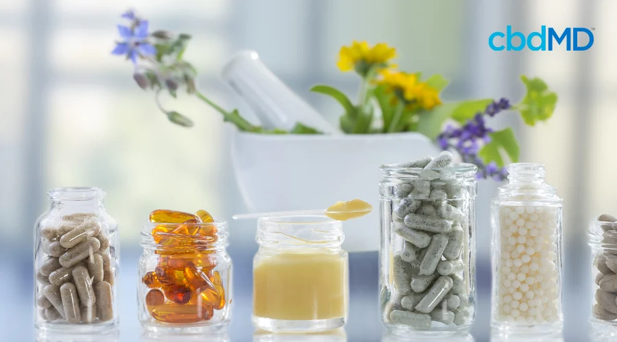 six jars filled with capsules, softgels, lotion, bead balls with mortar and pestle in background filled with variety of yellow, purple, blue flowers
