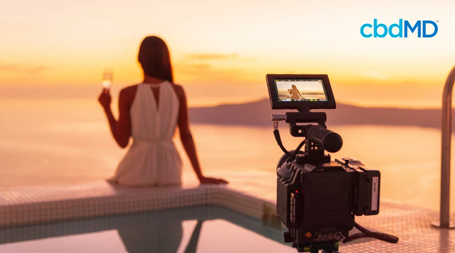 Dark-haired woman sits on the edge of a pool with a glass of wine in the background with video camera resting in the foreground