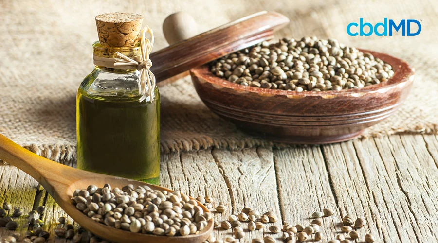 ooden spoon with seeds rests on wood table in front of bottle of oil with cork top and ceramic bowl with lid off filled with seeds sitting on cloth