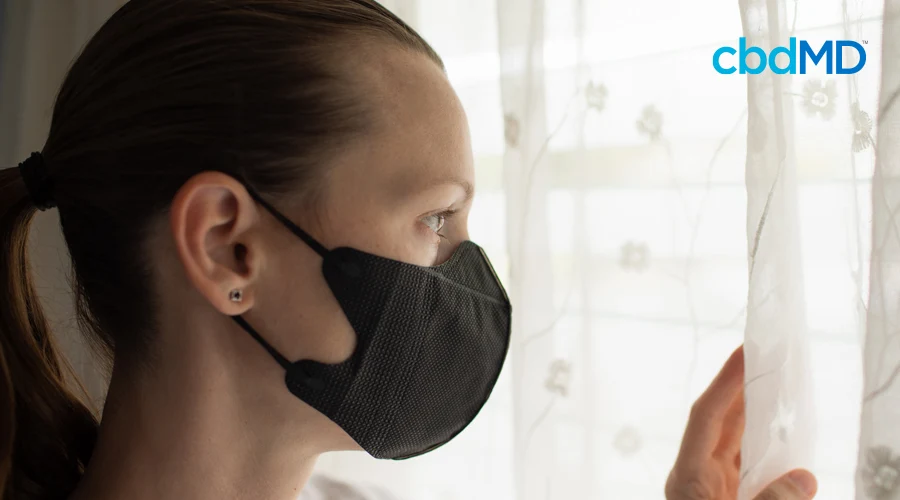 ark-haired woman in ponytail looks out of window with black face mask on