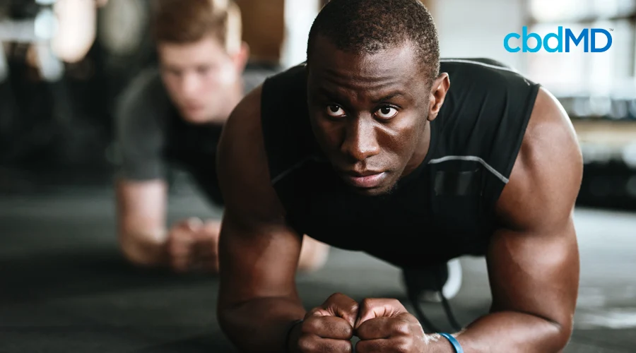 Man in black sleeveless workout shirt holding plank position with man in gray shirt doing plank in background at gym