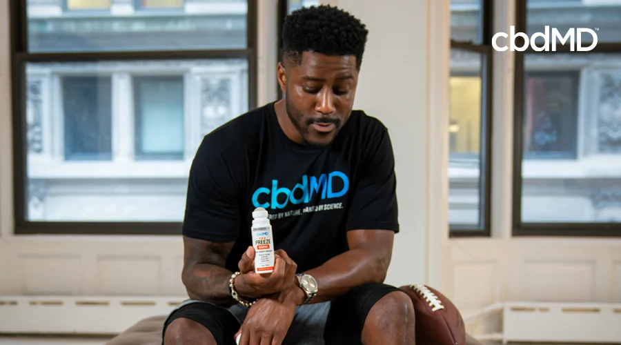 Team cbdMD Athlete and former football pro Nate Burleson wearing black cbdMD shirt showcasing roll-on bottle of 1500 mg CBD Freeze topical with cap removed and football at side