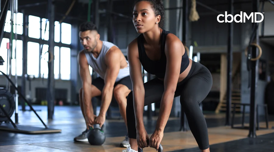 Dark-haired woman in black workout pants and tee squats with kettlebell in gym with man in gray shirt squatting with kettlebell in background