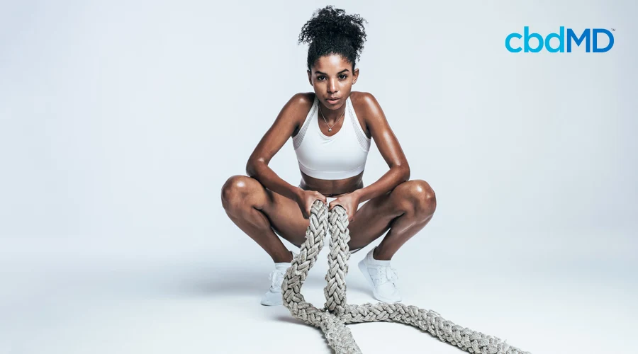 Woman with black curly hair in bun holds workout rope in squatting position wearing white workout top and shorts
