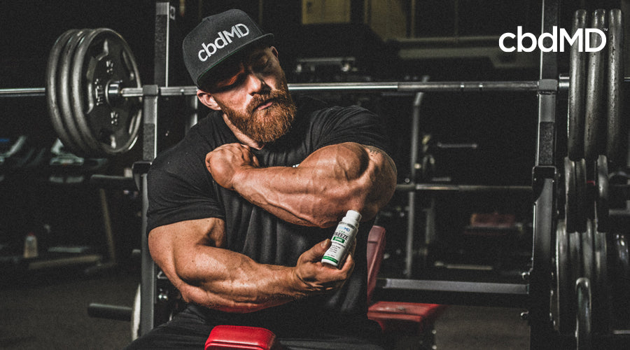 Bodybuilder James Flex Lewis applies 300 mg CBD Freeze roller to left arm wearing black cbdMD hat