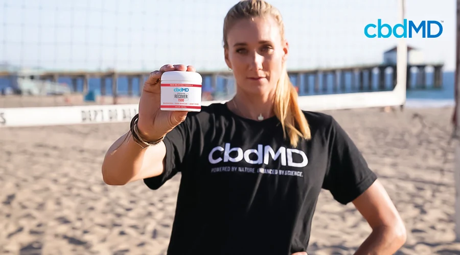 Kerri Walsh Jennings stands on beach in front of volleyball net holding cbdMD CBD Recover 1500 mg tub