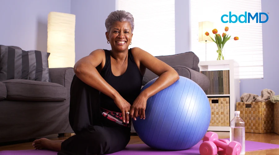 A woman sitting down on a yoga mat in her living room with a exercise ball weights and a water bottle