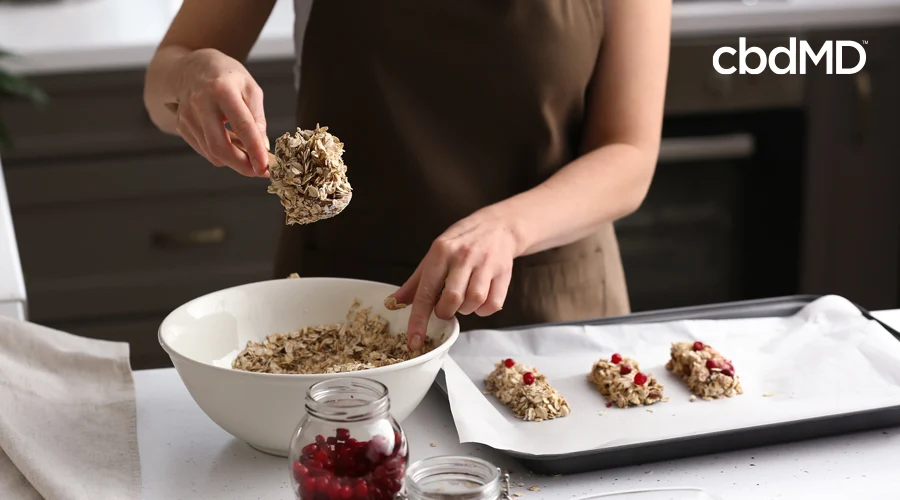 Woman in brown apron scoops protein balls of oats and berries out of mixing bowl onto baking sheet with parchment paper