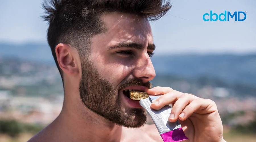 Shirtless dark-haired man with short hair and beard eats protein bar while standing outside