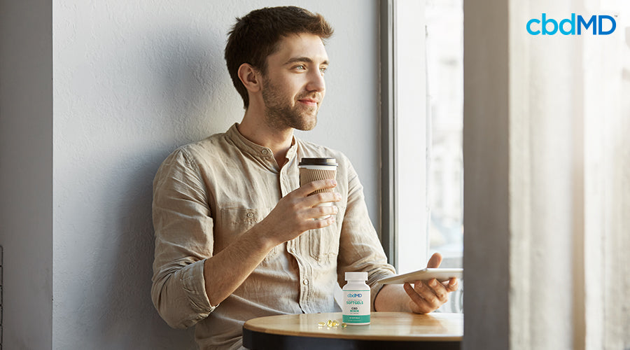 A man sitting down with a coffee and a tablet in his hands next to broad spectrum softgels - cbdMD
