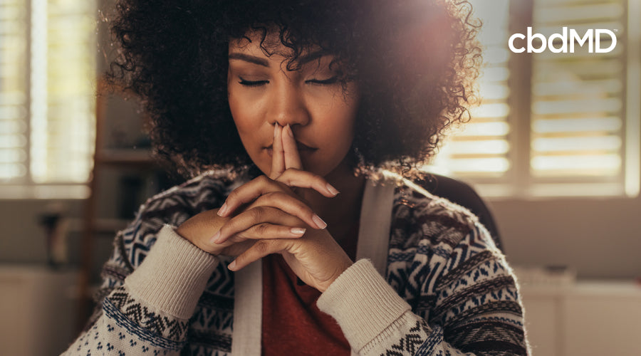A dark skinned woman with natural hair sits with her eyes closed and her fingers steepled in front of her lips