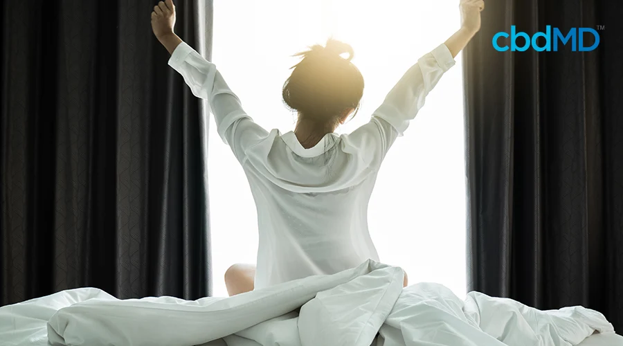 A woman in an oversized white shirt wakes up and stretches in front of dark curtains