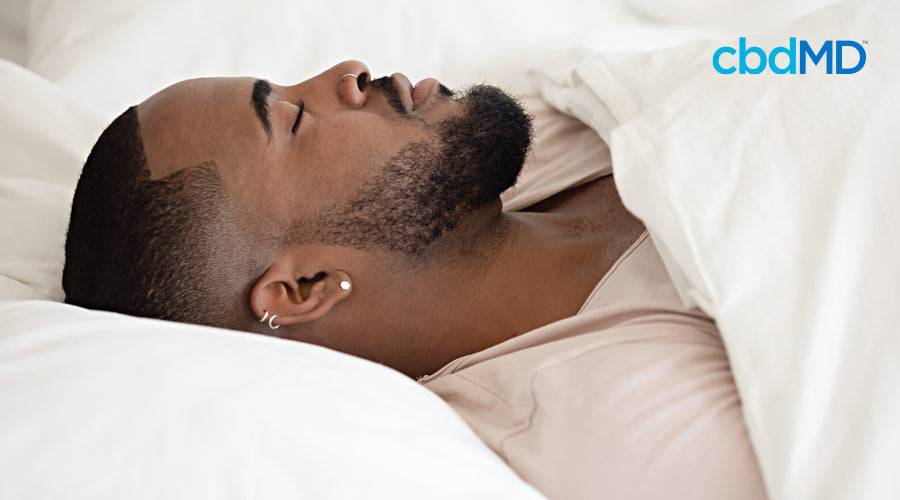 A dark skinned man with a trimmed beard sleeps soundly beneath fluffy white blankets