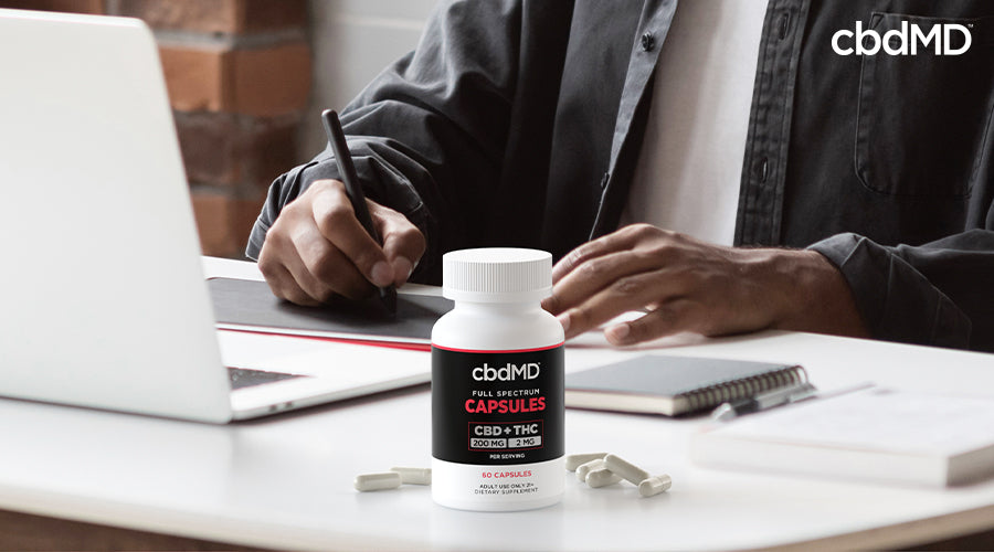 A man sitting down at a desk with a computer as he works with full spectrum capsules 200 mg cbd 2 mg thc in front of him - cbdMD