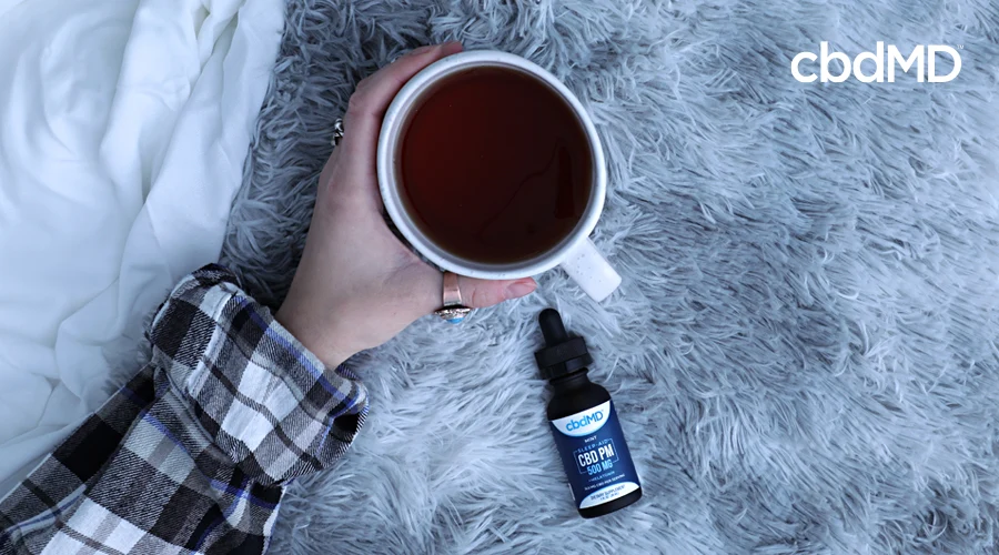 A bottle of cbd pm lays on a table next to an arm that is holding a cup of coffee