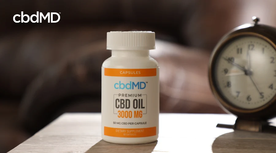 A bottle of 3000 mg cbdMD CBD Oil Capsules sits on a table next to an old alarm clock