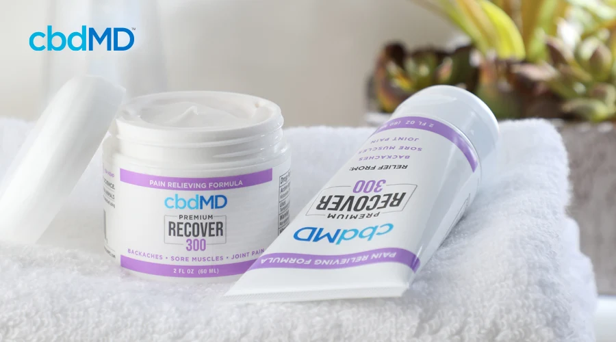 cbdMD's 300 mg CBD Recover tub rests on white towel with top unscrewed sitting next to squeeze bottle of 300 mg CBD Recover laying flat on towel