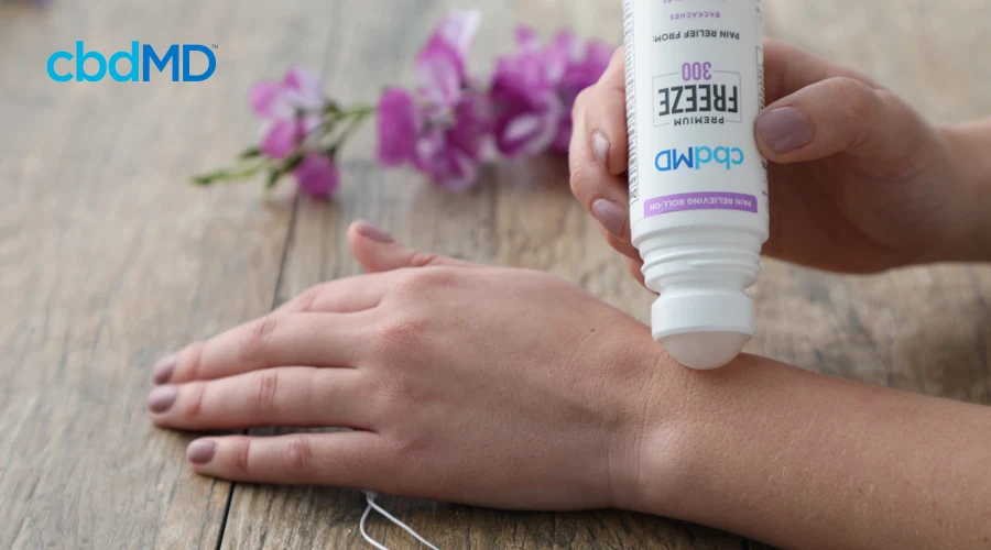 Hand rests on wood table applying 300 mg CBD Freeze roller by cbdMD with pink flower in background