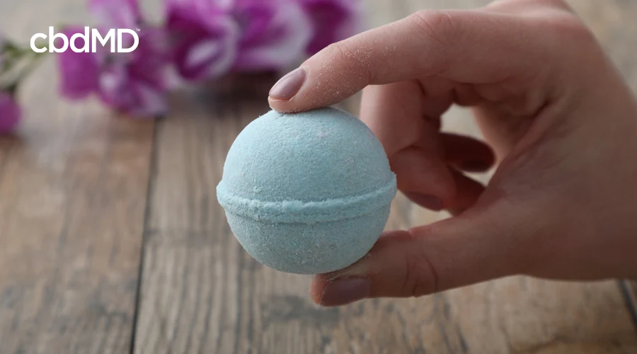 Hand holds blue Rejuvenate CBD bath bomb by cbdMD with pink flower in background