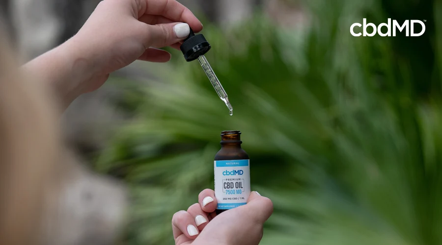 Manicured hands remove dropper with oil in it from cbdMD 7500 mg CBD oil tincture bottle with green plants in background