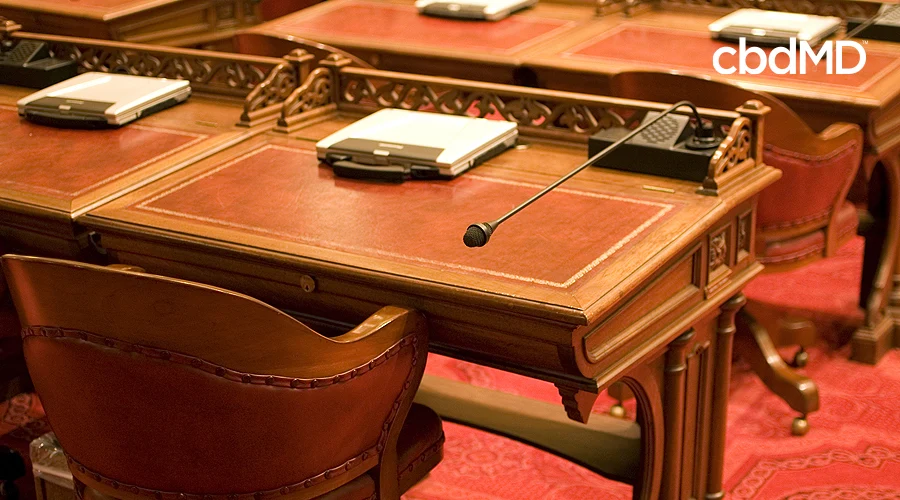 A seat in a legislative chamber sits empty with a microphone pointing back and a laptop sitting on the desk