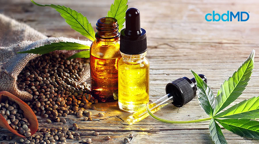Two bottles of cbd oil, one of them open, sit next to a full dropper beside hemp seeds and a hemp leaf