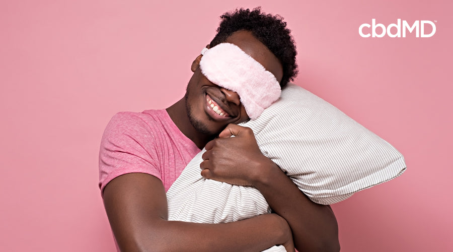 A dark skinned man hugs a pillow while wearing a pink sleep mask