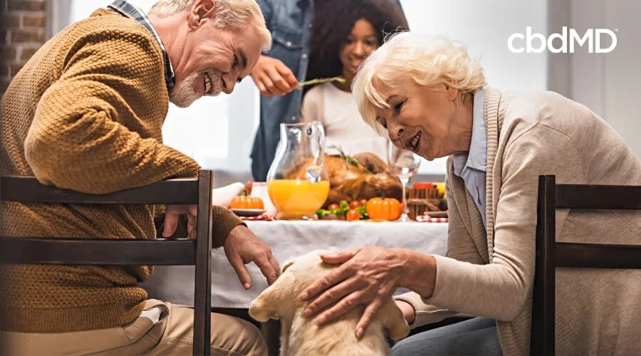 Two older people sit at a kitchen table and pet a yellow lab