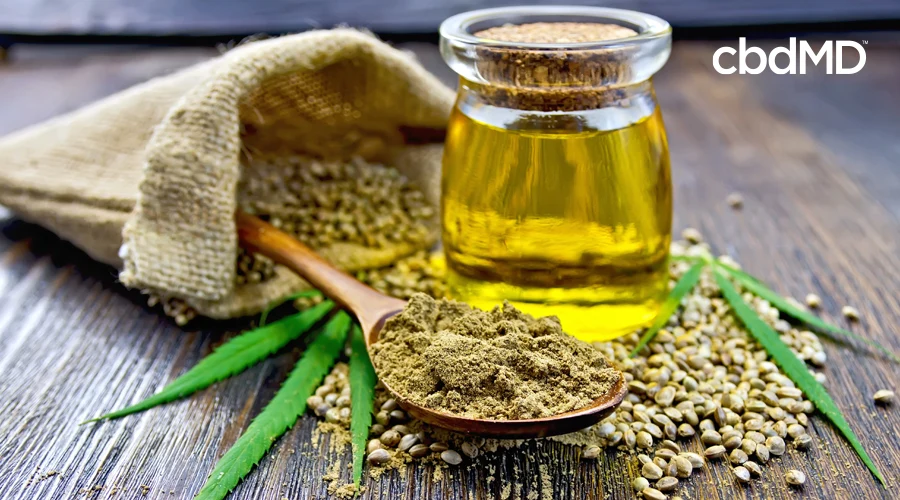 A glass jar of cbd oil sits among hemp seeds next to a wooden spoon