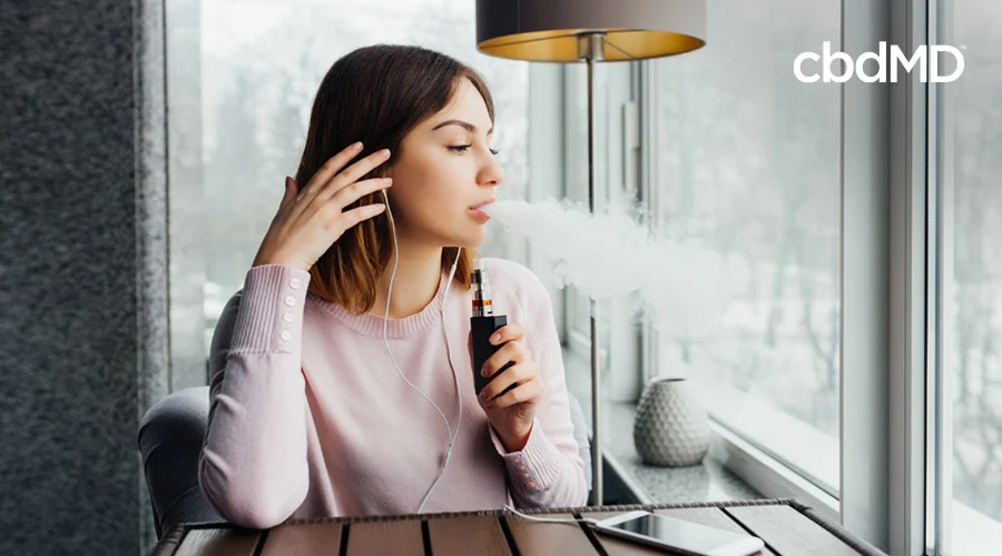 A dark haired woman sits in a coffee shop and blows out a cloud of vape smoke