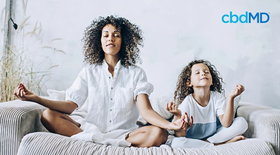 A dark skinned woman with natural hair sits cross legged on the couch next to her child