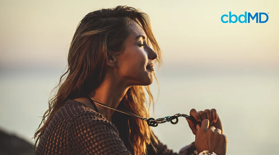 An attractive woman sits on the beach with the sun setting holding out a necklace away from her chest