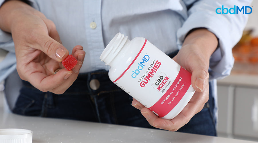 A woman holding a cbd gummy with her fingers and holding broad spectrum gummies 200 mg per serving tropical mix in her hand - cbdMD