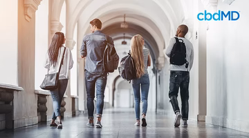 Four students with backpacks walking in a bright college hallway, cbdMD logo visible.