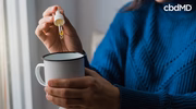 Person adding CBD oil to coffee mug with dropper, close-up view indoors