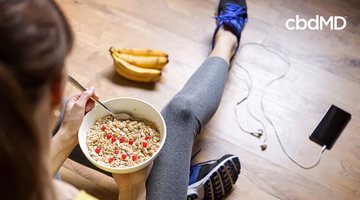 Woman in workout clothes eating healthy cereal with fruit, bananas and phone nearby on wood floor