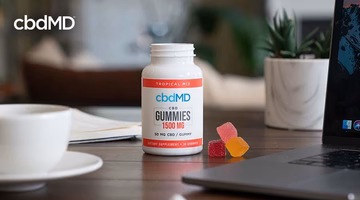 CBD gummies bottle on table with assorted gummies, coffee cup, laptop, and houseplants.