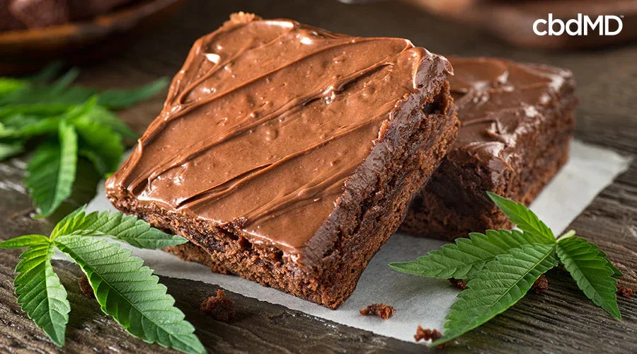 Frosted chocolate brownies with green hemp leaves on a rustic wooden surface.