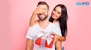 Smiling woman surprises man with Valentine's gift box, pink background, cbdMD logo visible